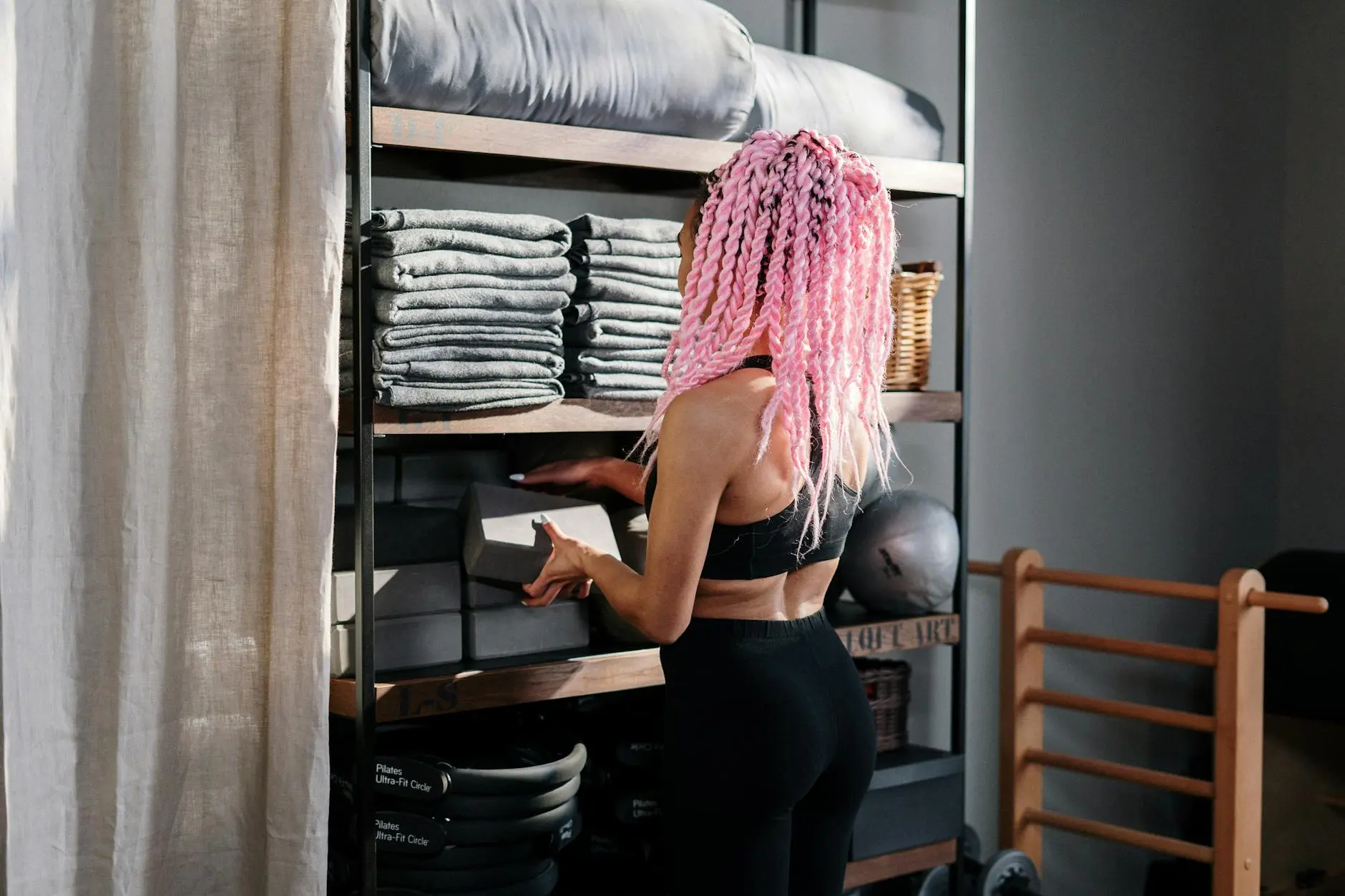A yoga instructor with pink braids arranges equipment in an indoor studio, promoting wellness and fitness.